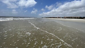 Une plage avec un ciel dégagé et des vagues. On aperçoit des bâtiments à l’arrière-plan.