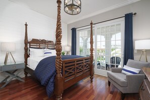 A four-poster bed with a wooden frame and a blue bedspread, a gray armchair with a blue pillow, and a glass desk with a lamp.