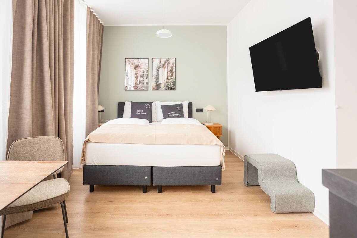 Quarto de hotel moderno com cama, escrivaninha, um banco e TV fixada na parede.