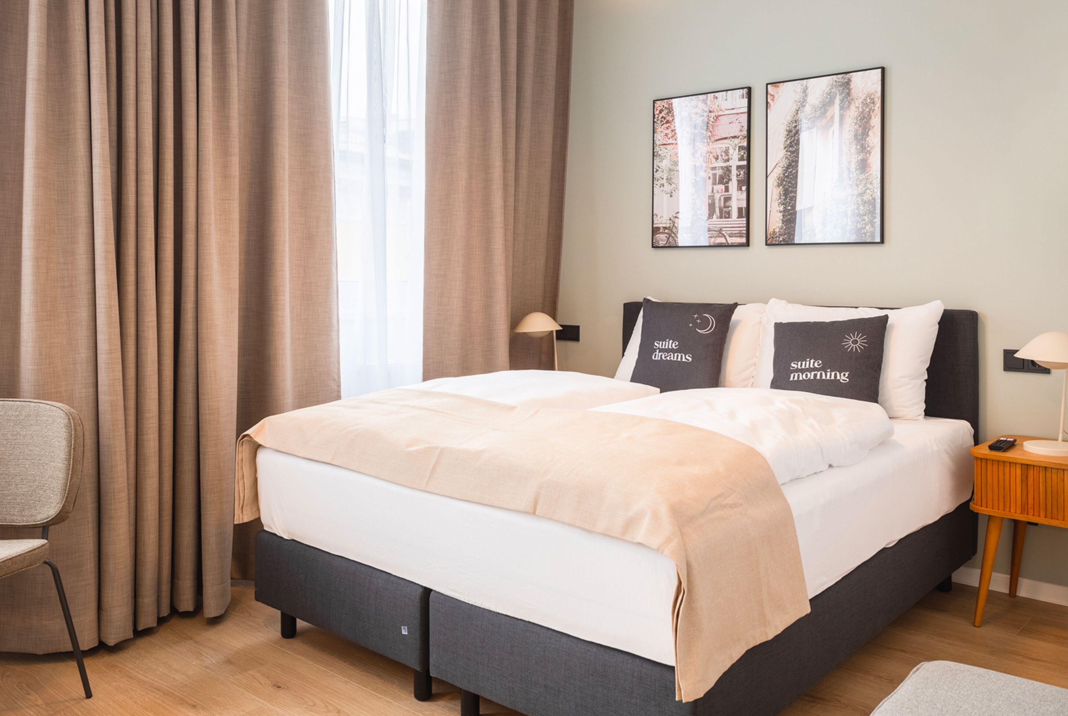 Una cama bien hecha con una manta color beige, una silla y dos lámparas de pared.