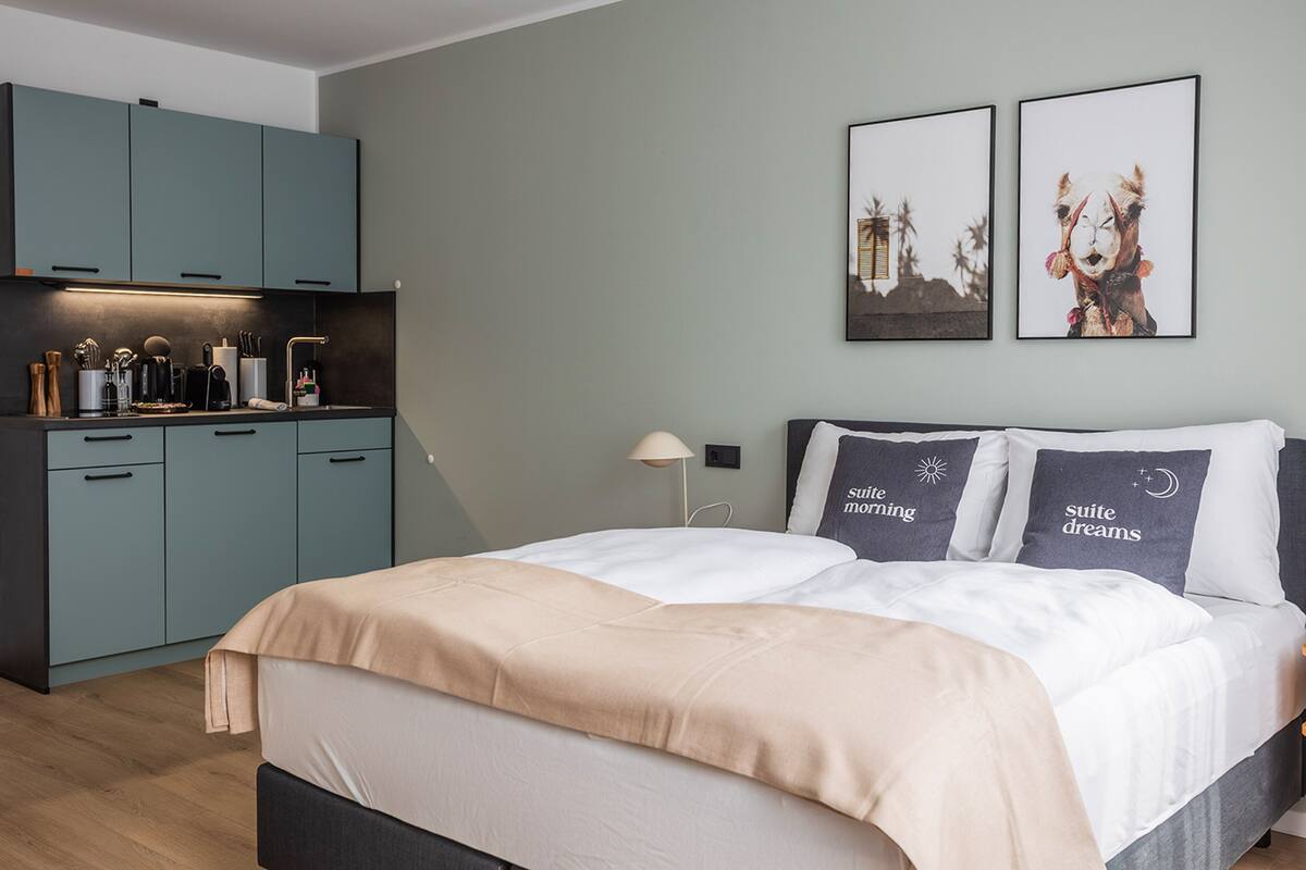 Quarto moderno com cama, mesas de cabeceira, um criado-mudo, luminária e uma área de kitchenette com armários e bancada.