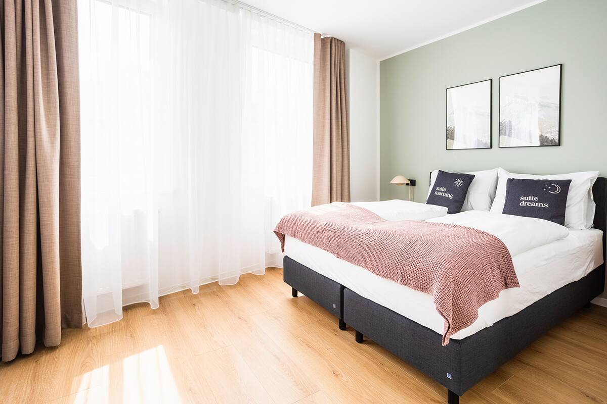 Quarto com cama, cortinas, mesa de cabeceira e duas imagens emolduradas na parede.