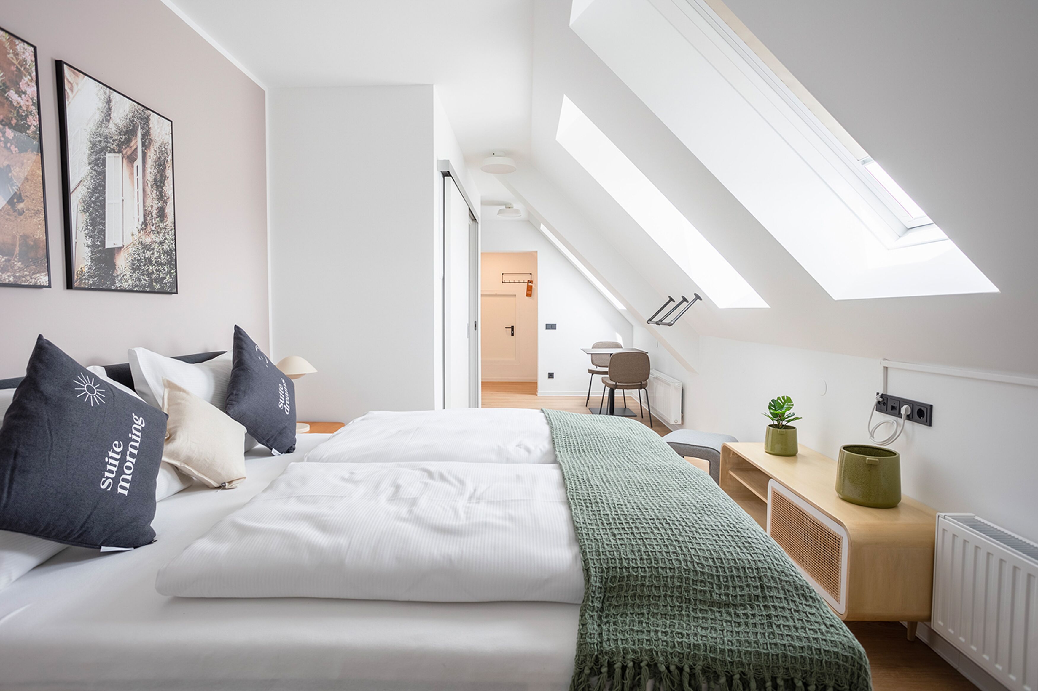 Een moderne slaapkamer met dakraam, een bed met witte lakens, een groene plaid en een kleine plant op een nachtkastje.