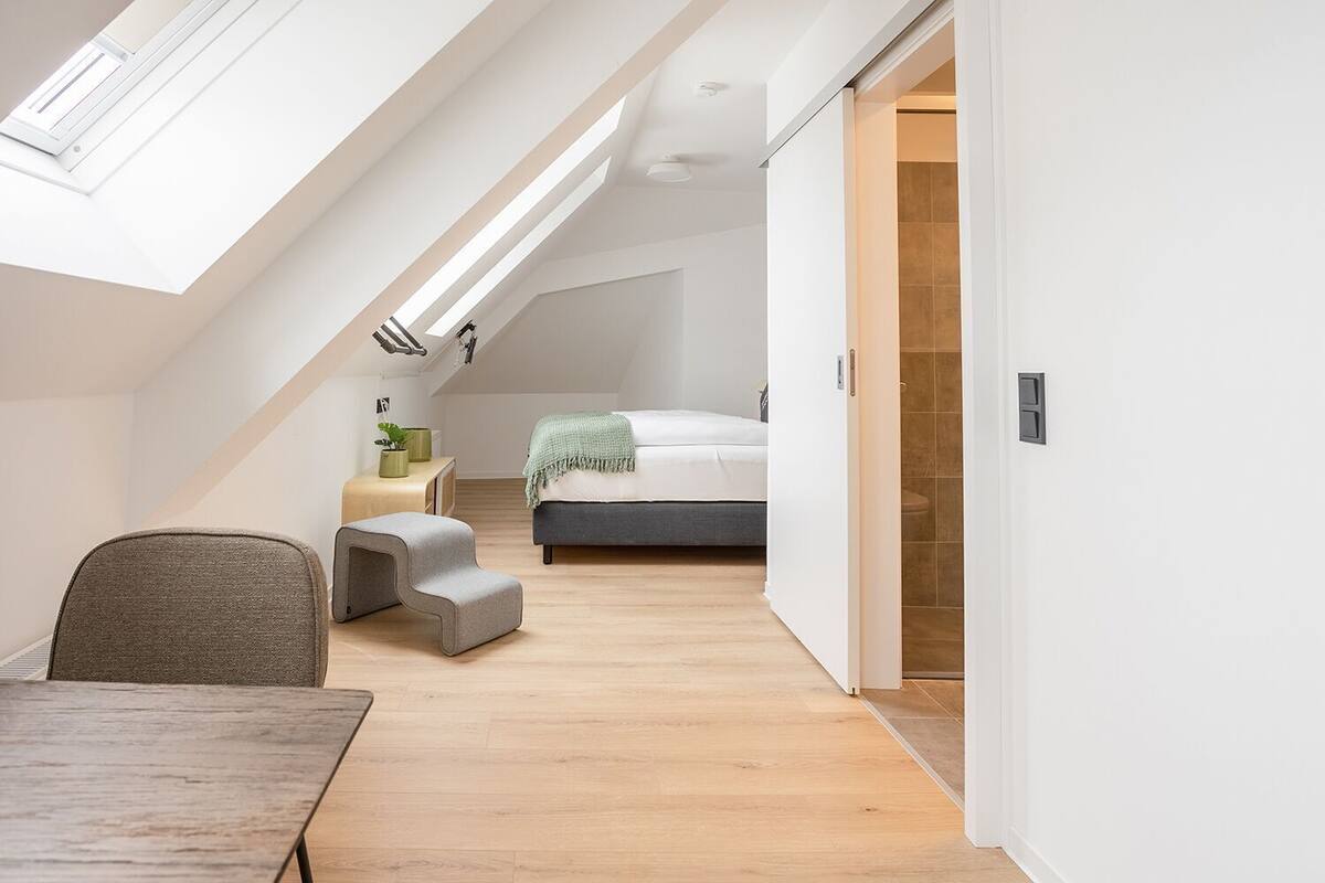 Quarto moderno no estilo loft, com janela no teto, uma cama, um pequeno espaço de estar e vista para o banheiro.