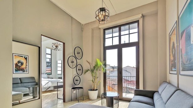 A modern living room with a grey sofa, a potted plant, and a large window with a view of buildings. - w* | Premium 1BR in Casco Antiguo (Panamá)