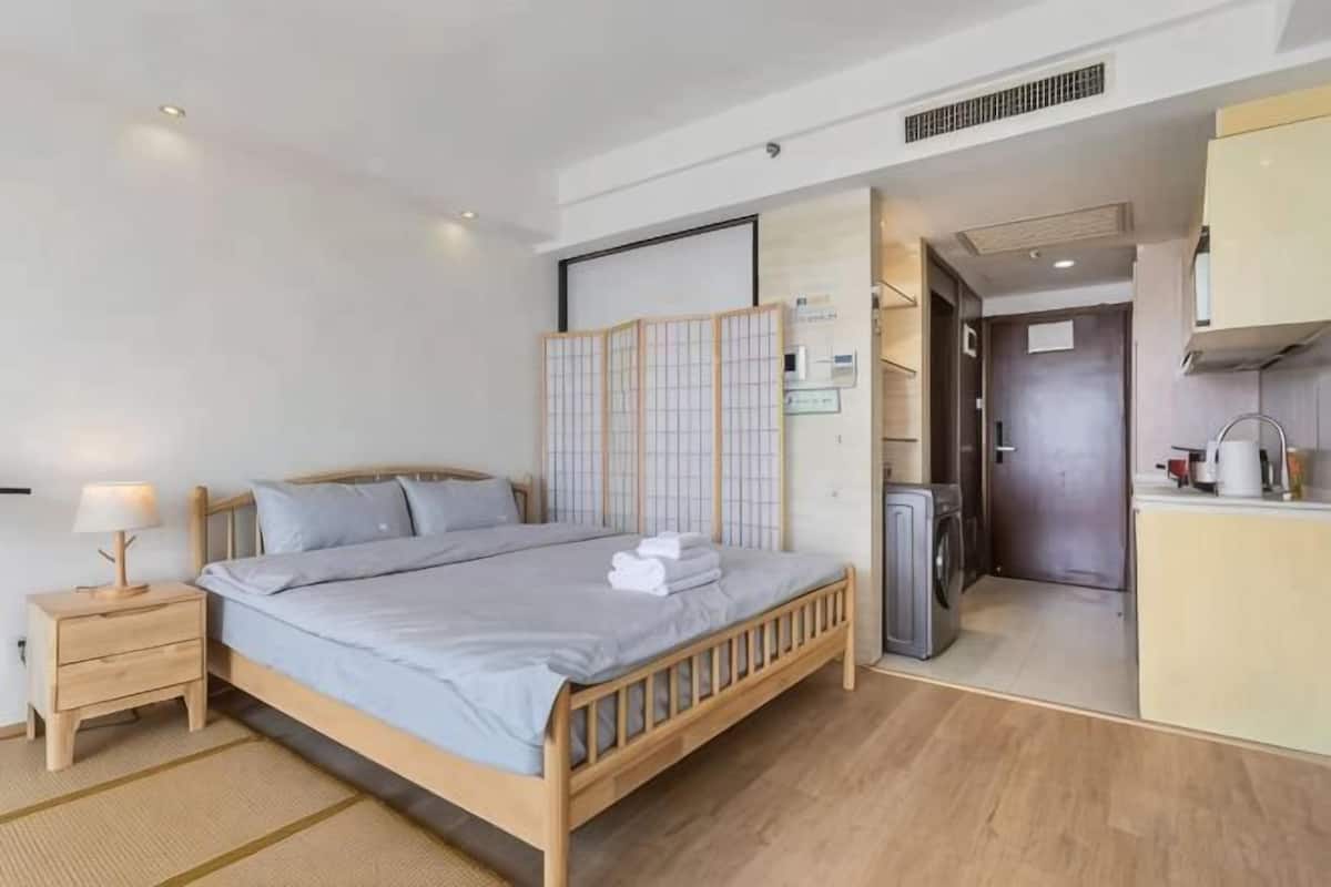 A bedroom with a bed, bedside table, and a view into a kitchen area.