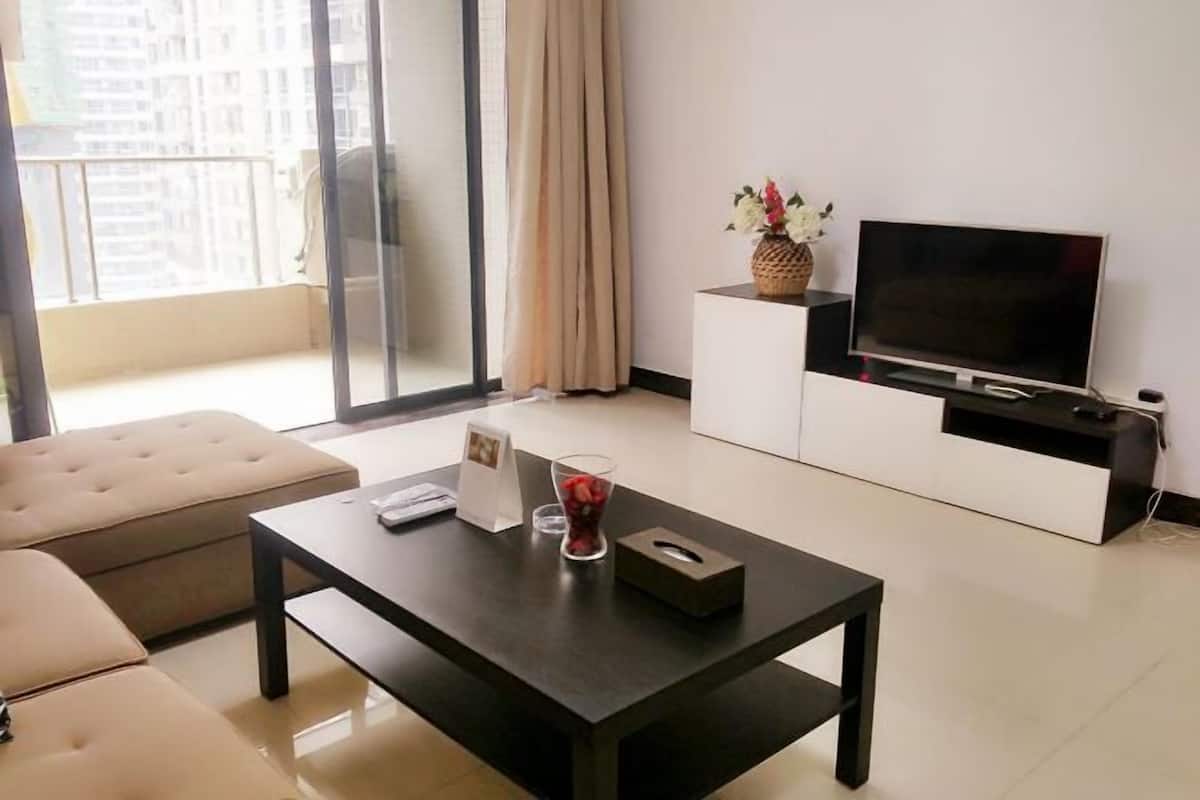A modern living room with a sofa, a coffee table, a TV on a white cabinet, and a balcony with a view.