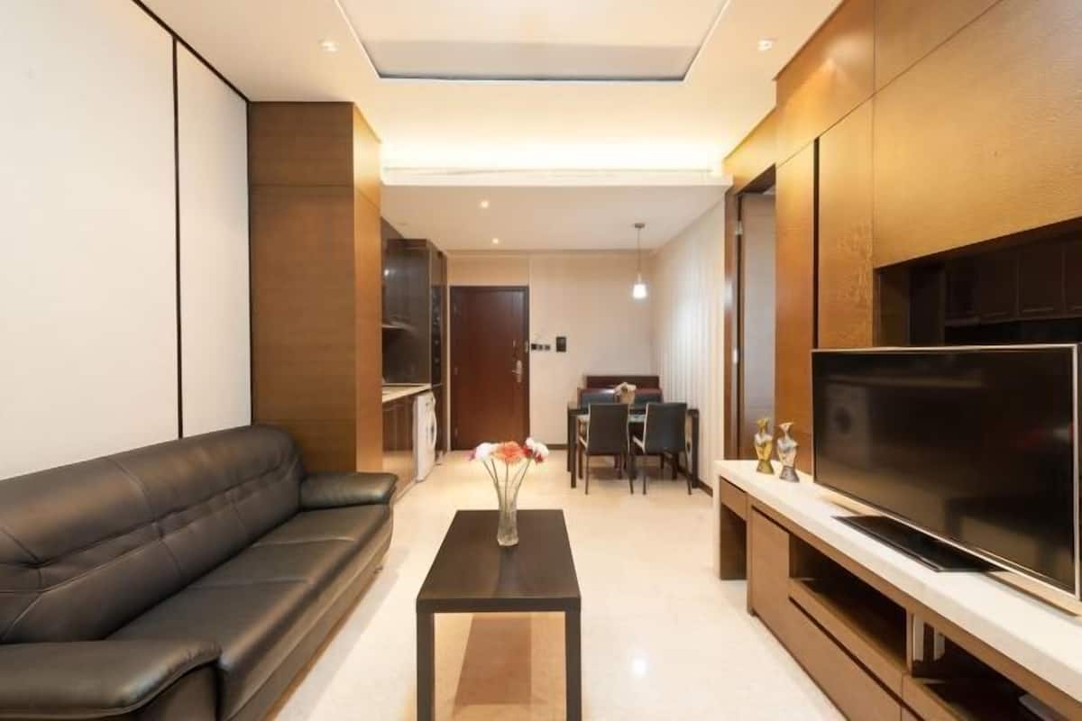 A modern living room with a black leather sofa, a coffee table with a vase, a flat-screen TV, and a dining area in the background.
