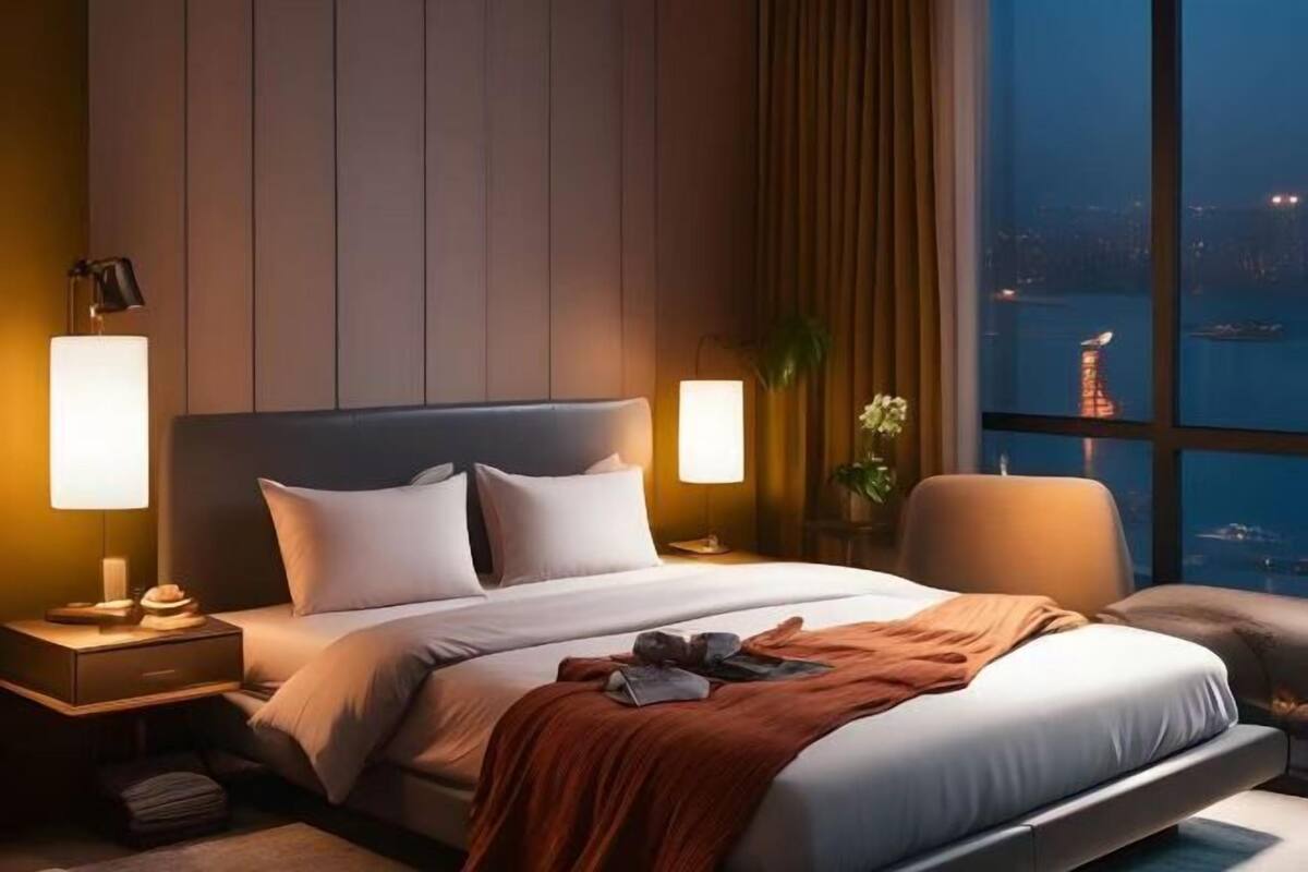 A modern hotel room with a large bed, two bedside lamps, a nightstand, and a view of a cityscape through large windows.