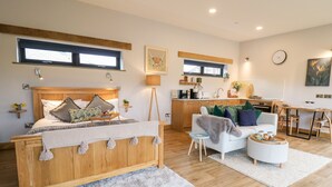 A modern bedroom with a wooden bed, a bedside table with a lamp, and a view of the kitchen area. - Tin Shed (Rhayader)