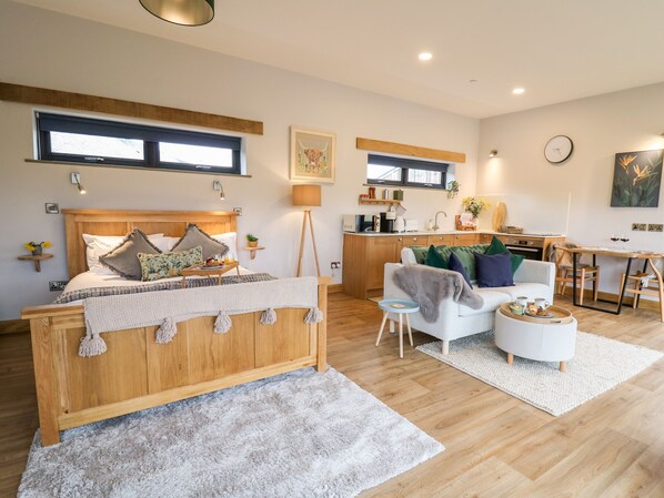 Quarto moderno com cama de madeira, mesa de cabeceira com abajur, e vista para a área da cozinha.