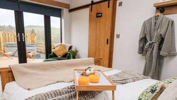 A hotel room with a bed, a wooden bedside table with a tray of breakfast items, a green chair, a wooden wardrobe, and a view of the outdoors. - Tin Shed (Rhayader)