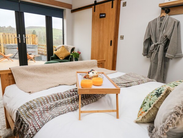Ein Hotelzimmer mit einem Bett, einem Nachttisch aus Holz mit einer Auswahl an Frühstücksgegenständen, einem grünen Sessel, einem Holzschrank und Blick ins Freie.
