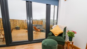 A room with a large sliding glass door leading to an outdoor area with a wooden deck and a chair. - Tin Shed (Rhayader)