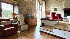 A rustic room with stone walls, a fireplace, a wooden chair, a desk, and a large window.