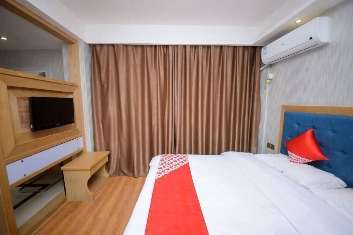 A hotel room with a bed, a TV on a wooden stand, a small table, a red and white patterned bedspread, a blue headboard, and a brown curtain.