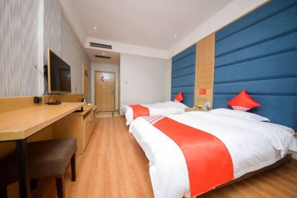 A hotel room with a wooden desk, two beds with red pillows, a TV, and a blue headboard.