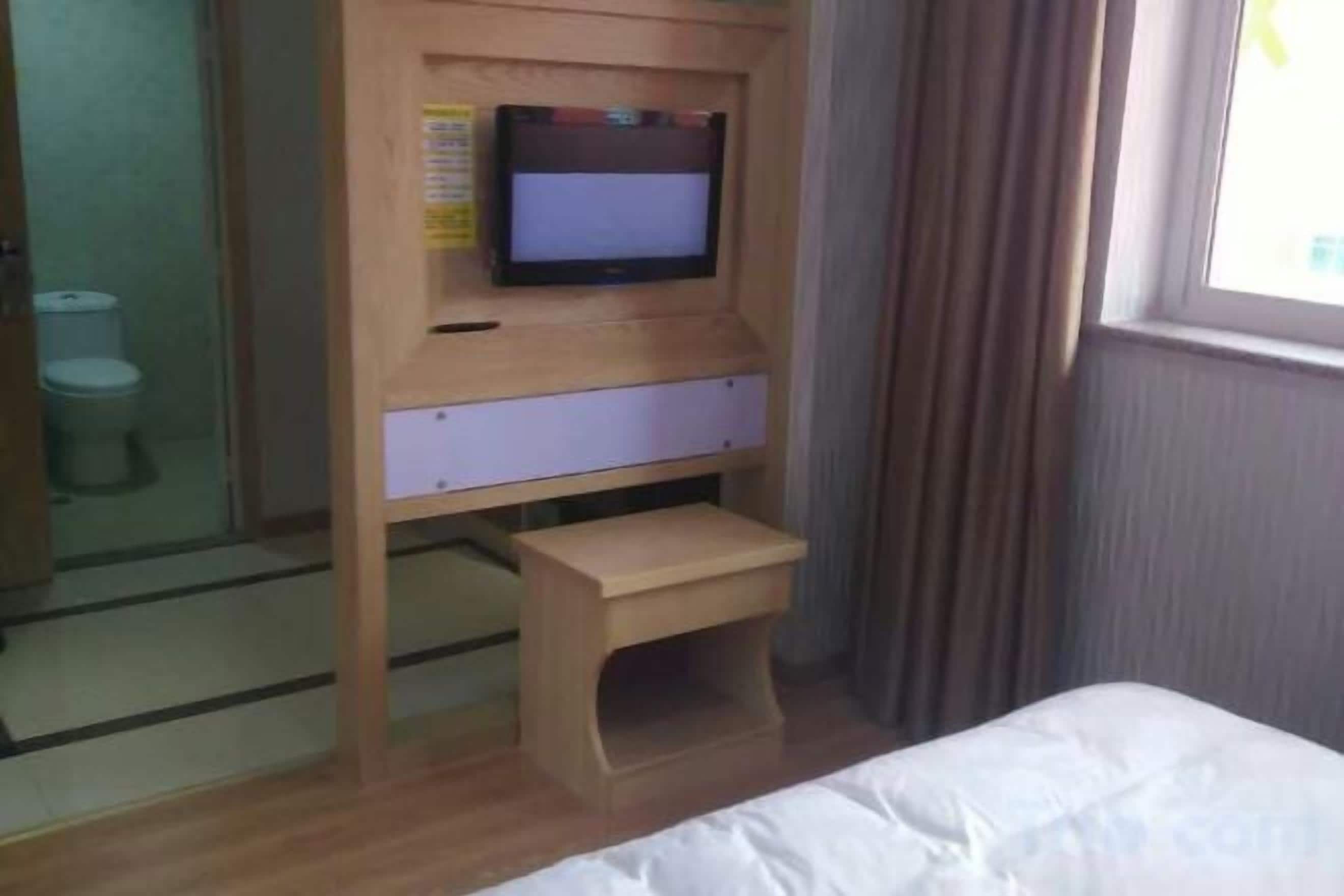 A hotel room with a bed, a wooden desk with a television, a small wooden stool, and a window with curtains.