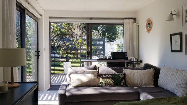 Un salon confortable avec un canapé, une table basse et un téléviseur. Une porte-fenêtre coulissante donne accès à un balcon agrémenté de plantes et d’une clôture.