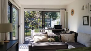 Ein gemütliches Wohnzimmer mit Sofa, Couchtisch und Fernseher. Eine Glastür öffnet sich zu einem Balkon mit Pflanzen und einer Umzäunung.