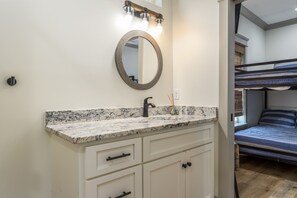 A bathroom with a marble countertop, a round mirror, and a sink. There is a bunk bed visible in the adjacent room.