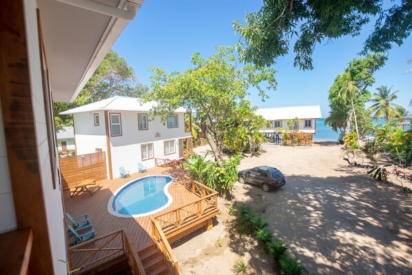A pool area with a wooden deck, a car parked nearby, and a view of a white building with multiple windows. - Casa Palmera 2 Bedroom Home by RedAwning (Roatan)