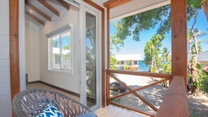 A balcony with a wicker chair and a wooden bench, overlooking a beach with palm trees and a building in the background. - Casa Palmera 2 Bedroom Home by RedAwning (Roatan)
