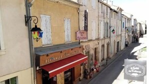 A narrow street with buildings on either side. A bakery with a red awning and a sign that reads "Boulangerie" is visible. There are hanging signs above the street and a lamppost on the left. - Aigues-Mortaise house in the city (Aigues-Mortes)
