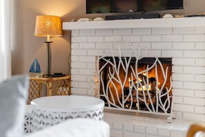 A living room with a white brick fireplace, a television mounted above it, and a round table with a patterned tablecloth.