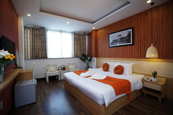 Quarto de hotel com uma cama grande, mesa de cabeceira, escrivaninha, cadeira, pequena geladeira e janela com cortinas.