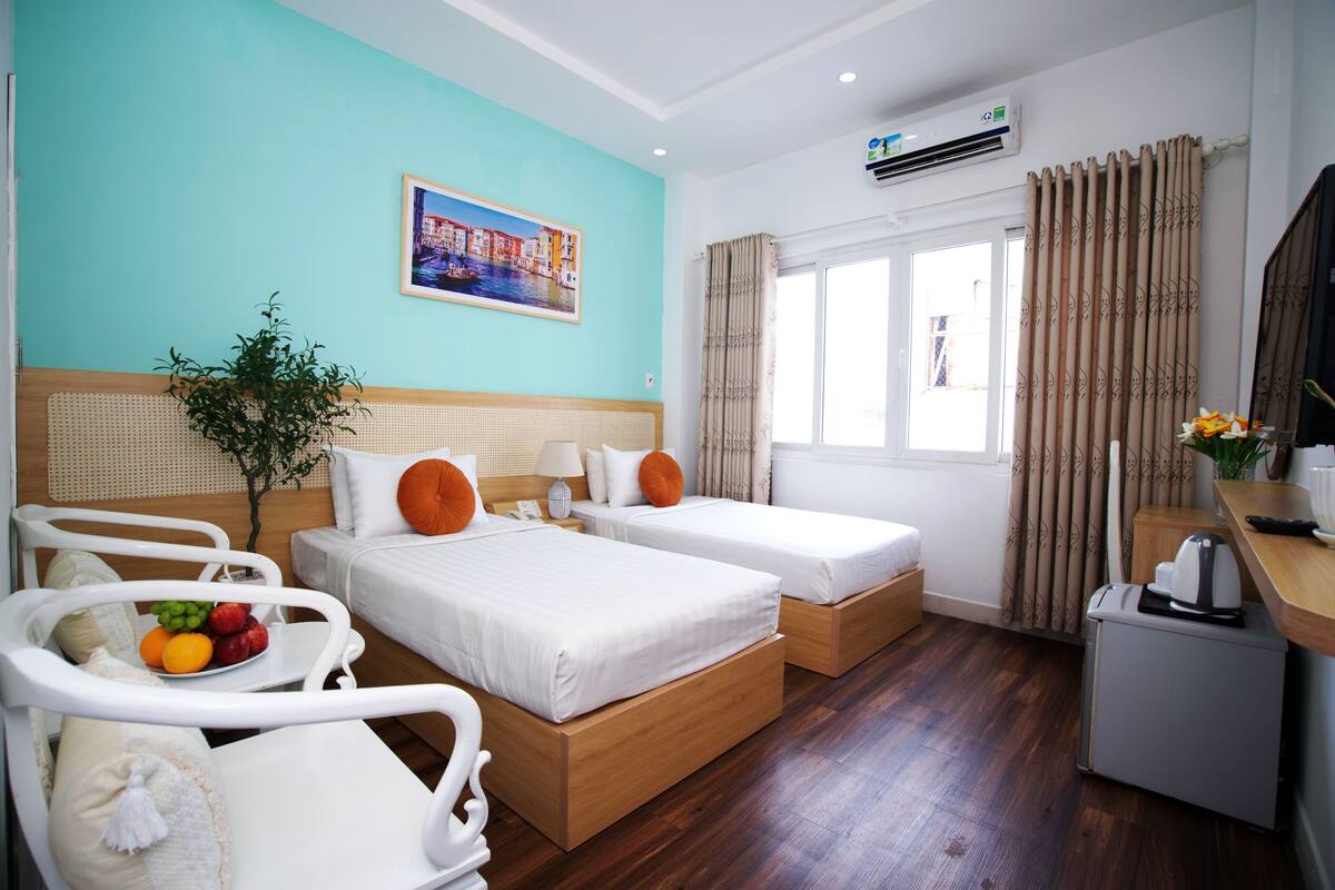 A hotel room with two beds, a small desk, a plant, and a window with curtains.