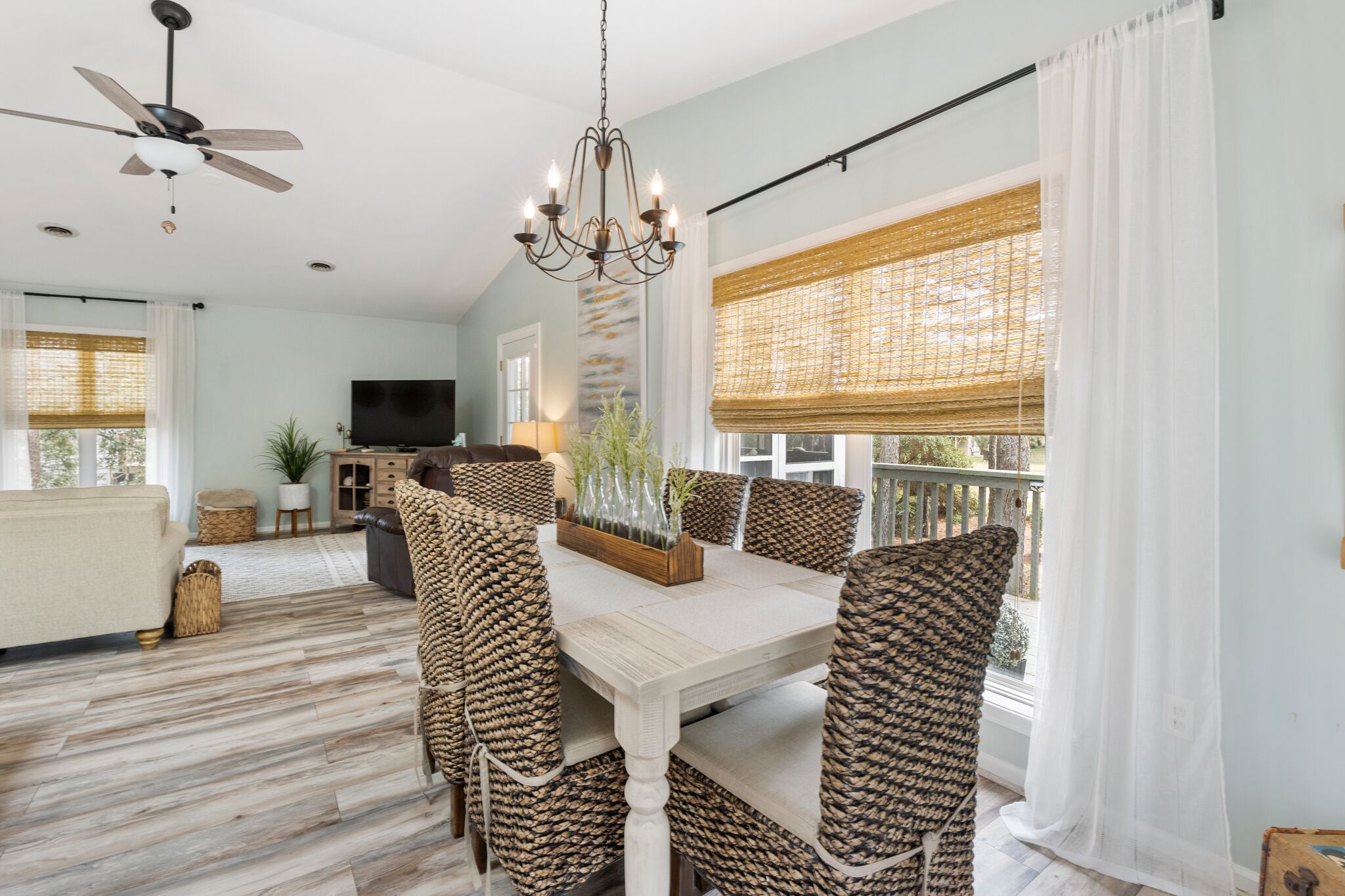 A dining area with a white table and wicker chairs, a chandelier, and a television in the background.