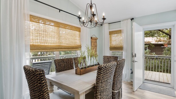 Uma área de refeições com mesa de madeira e cadeiras de fibra, um lustre e janelas amplas com persianas de bambu.