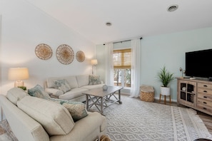A living room with a sofa, coffee table, and TV stand. There are decorative wall pieces and a window with curtains.