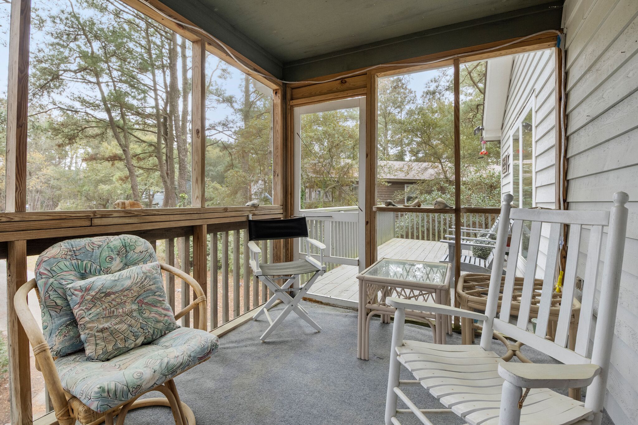 A covered porch with wicker chairs, a small table, and a view of trees and neighboring houses.