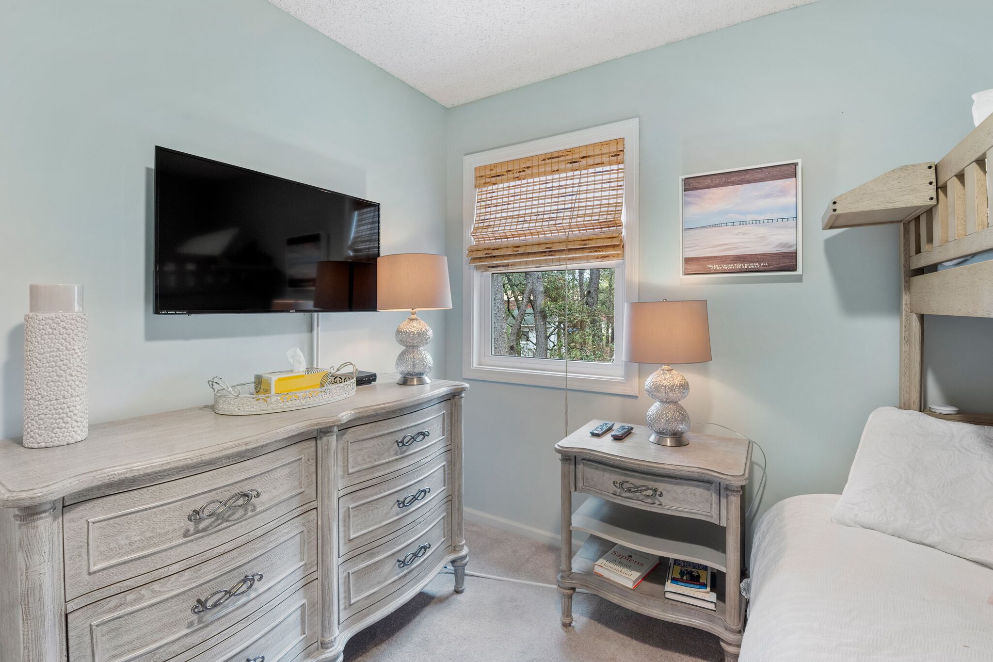 A bedroom with a TV, two bedside tables with lamps, a dresser, and a window with blinds.