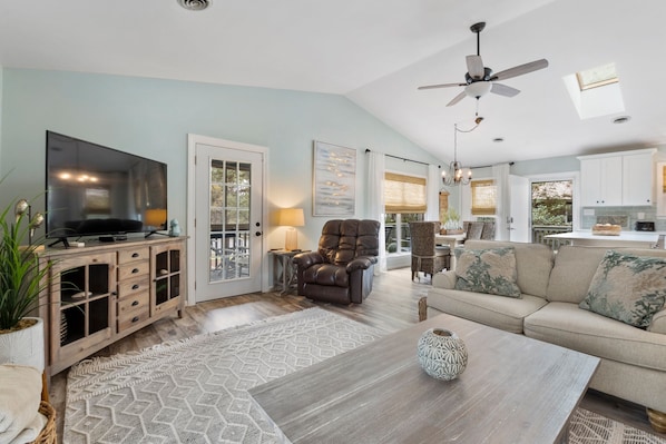 A spacious living room with a large flat-screen TV, a comfortable sofa, a leather armchair, and a dining area with a kitchen in the background.