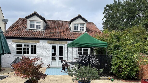 A house with a tiled roof, a white exterior, and a green door. There is a garden with a pool, potted plants, and a patio area. - The Lodge, South Cotswolds, contemporary rural home, Spa Pool, views, parking (Chippenham)