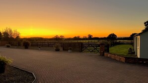 A paved driveway leading to a gated entrance with a wooden fence, surrounded by greenery and a building with a white exterior. - The Lodge, South Cotswolds, contemporary rural home, Spa Pool, views, parking (Chippenham)