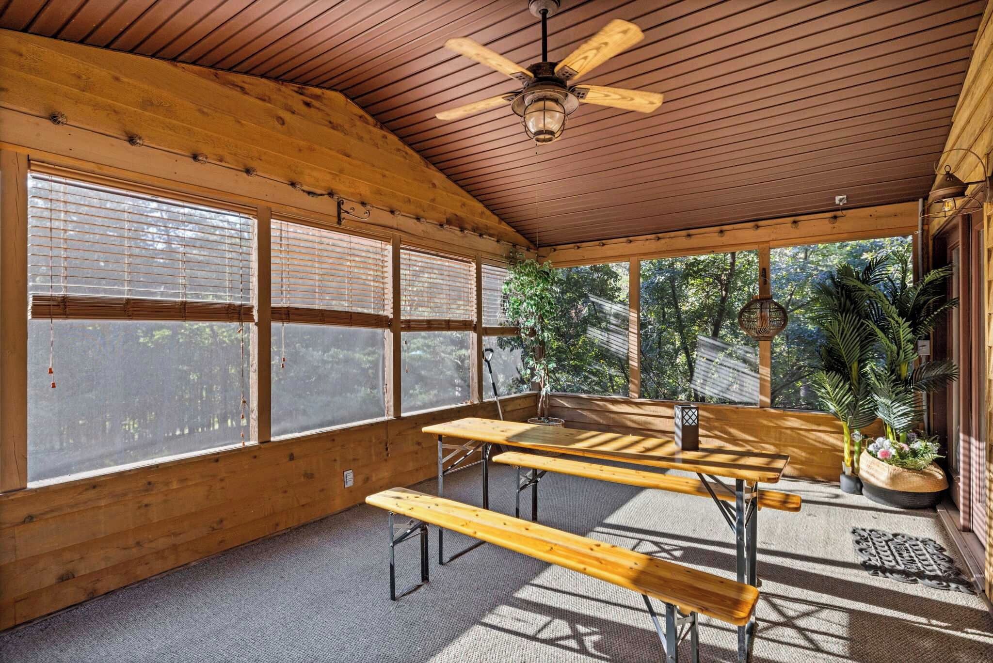 A covered outdoor seating area with wooden tables and benches, large windows, and a ceiling fan.