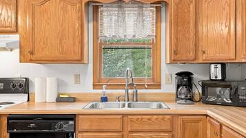 A kitchen with wooden cabinets, a double sink, a window with lace curtains, and a coffee maker.