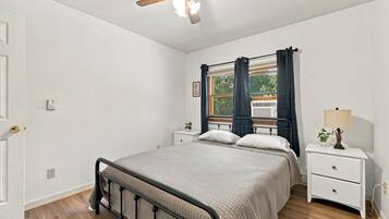 A bedroom with a bed, bedside tables, a ceiling fan, a window with curtains, and a wall-mounted air conditioner.
