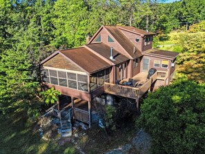 A multi-level wooden cabin with a deck, surrounded by dense forest. - Group-friendly cabin w/ hot tub & deck (Great Cacapon)