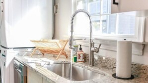 Une cuisine équipée d’un évier en acier inoxydable, d’un étendoir à linge en bois et d’une fenêtre donnant sur des immeubles.