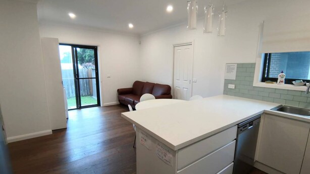 A modern kitchen with a white island, a brown leather sofa, and a sliding glass door leading to a backyard. - A Cozy room in Center of Glen (Glen Waverley)