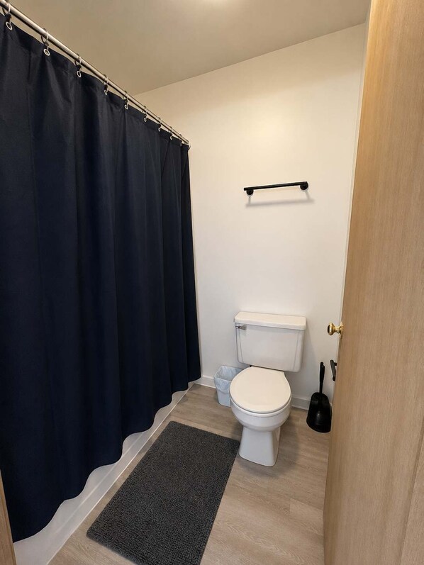 Un baño con un inodoro, una cortina de ducha, un tapete pequeño y un estante con un objeto negro.