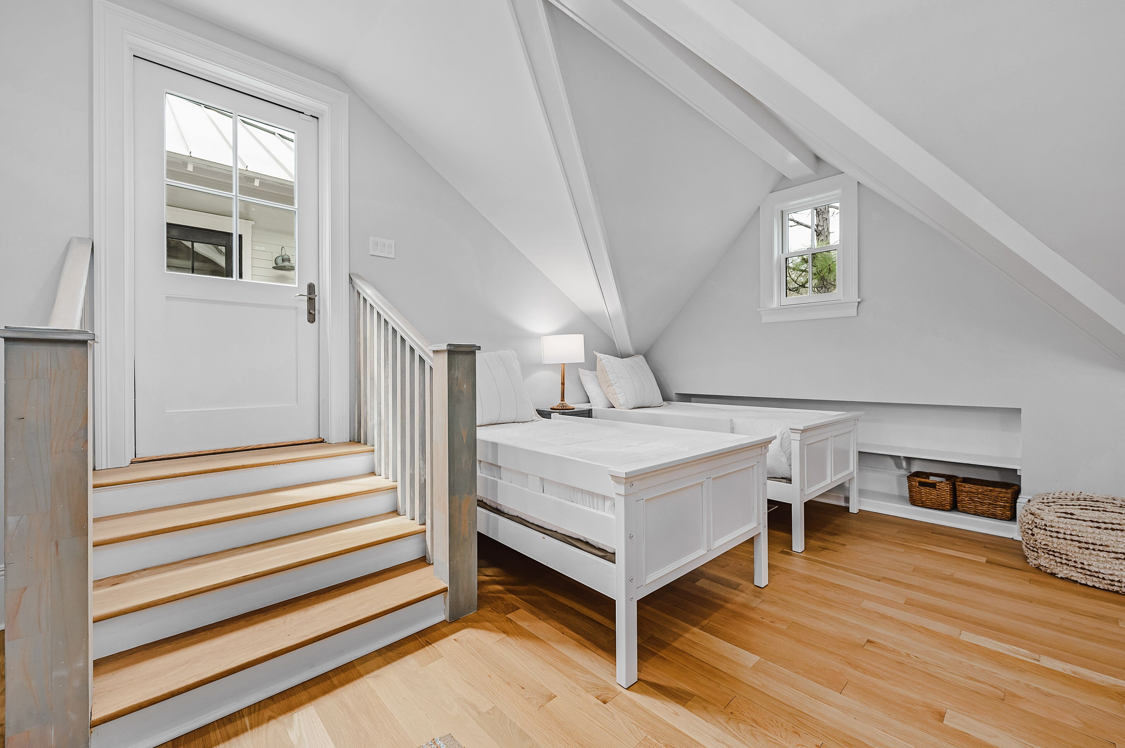 Une chambre à coucher avec un lit blanc, une table de chevet, une lampe, une fenêtre et un escalier.
