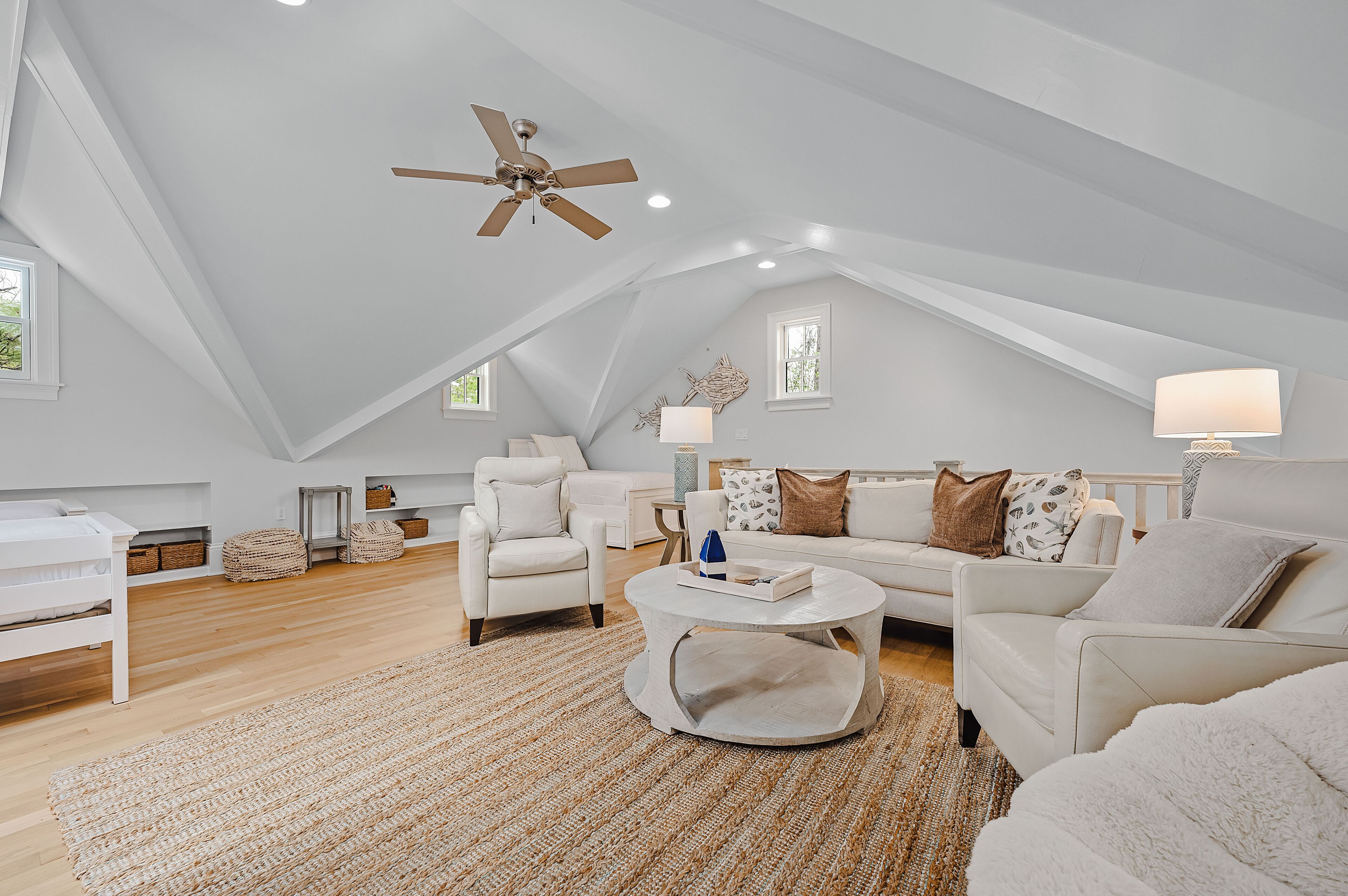 A spacious loft bedroom with a high ceiling, a ceiling fan, a large window, a white bed, a white sofa, a coffee table, and a rug.