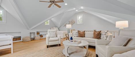A spacious loft bedroom with a high ceiling, a ceiling fan, a large window, a white bed, a white sofa, a coffee table, and a rug.