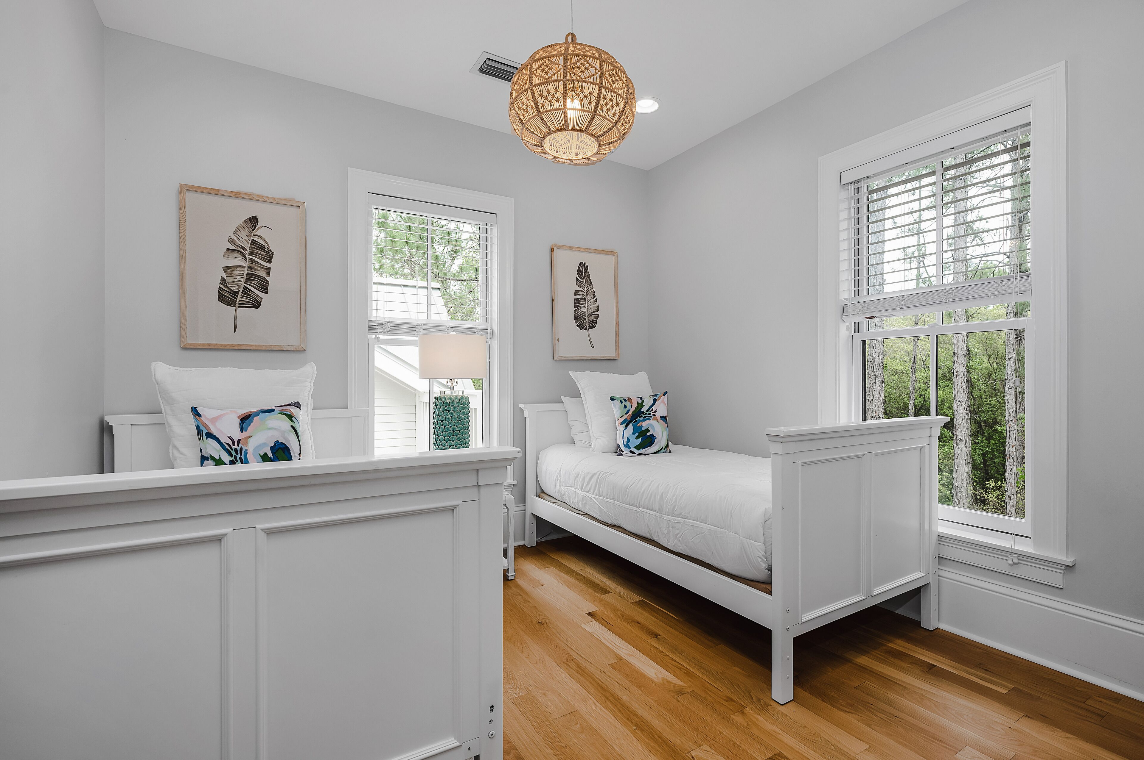 A bedroom with a white bed, two windows with blinds, a ceiling fan, and two framed leaf artworks.